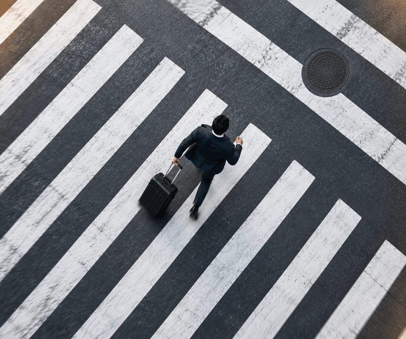 Overhead view of a young professional crossing a city road on a zebra crossing, pulling a rolling suitcase